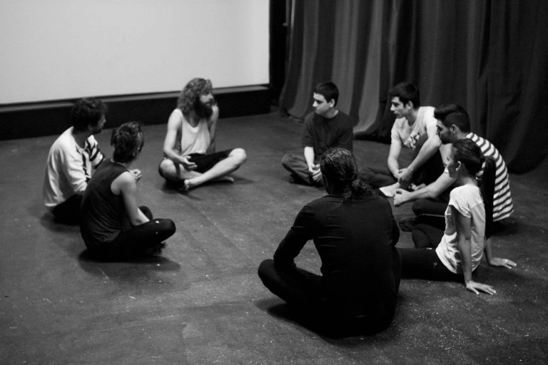 Actores y director sentados en el suelo del aula de teatro con la pantalla de cine de fondo y hablando del curso recibido como cierre de este