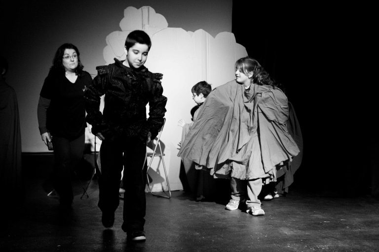 Alumnos y profesora en un ensayo en la sala de teatro con un fondo de escenografía