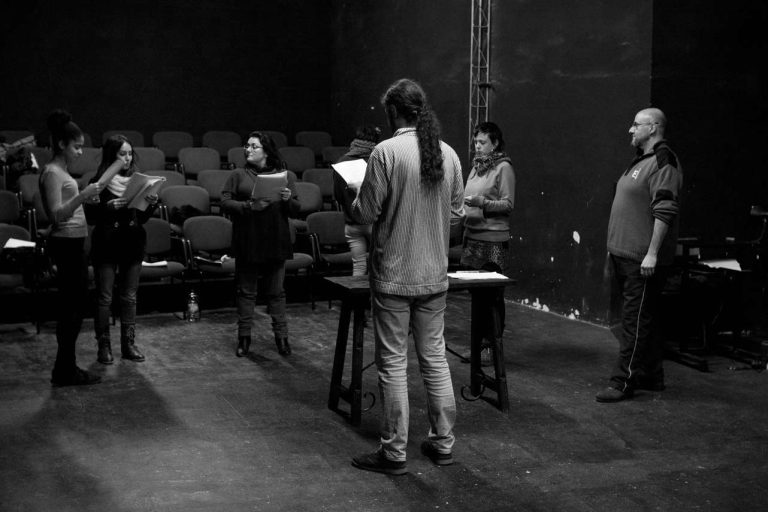 Grupo de actores y profesor en la sala de teatro con las butacas de fondo leyendo los textos teatrales