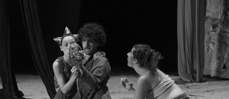 Actrices y actor en el escenario con telas aéreas colgando, fondo negro y escenario lleno de arena en el suelo. El actor central sostiene un botella ante la mirada atónita de las otras dos intérpretes