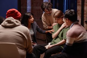 Grupo de actrices y actores en el aula. Dos chicos están de espaldas. Uno de ellos lleva una sudadera con capucha y un gorrito de lana roja, y el otro chico un suéter y una camisa. Otras dos chicas de frente, sentadas en sillas. Una de ellas está apoyando una de las chicas en actitud triste y con pañuelos blancos en una mano. Otro compañero pero de pie y apoyando con las manos como señal de ánimo