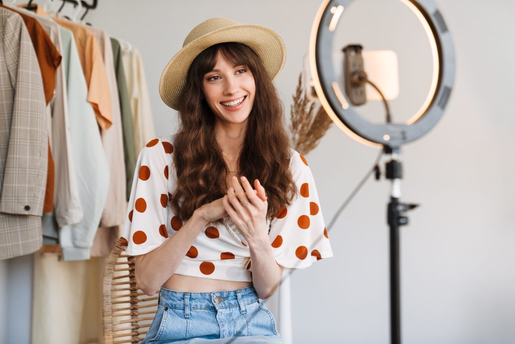 Chica con pantalón vaquero y camiseta corta con redondelitos rojos tipo flamenca y con sombrero de paja redondo en la cabeza. Está con el trípode una luz y el móvil haciéndose un selfie. Tiene detrás de ella camisas y chaquetas como parte del vestuario