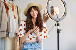 Chica con pantalón vaquero y camiseta corta con redondelitos rojos tipo flamenca y con sombrero de paja redondo en la cabeza. Está con el trípode una luz y el móvil haciéndose un selfie. Tiene detrás de ella camisas y chaquetas como parte del vestuario