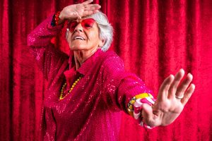 Mujer mayor de edad haciendo un gesto con las manos. Una de las manos con el brazo extendido hacia delante. La otra mano llevándola a la frente y el codo doblado. Lleva ropa muy colorida y con brillos. Colgantes y pulseras de colores. Está de plano americano y sobre un telón de fondo rojo. Lleva unas gafas de color rojo en forma de corazón.