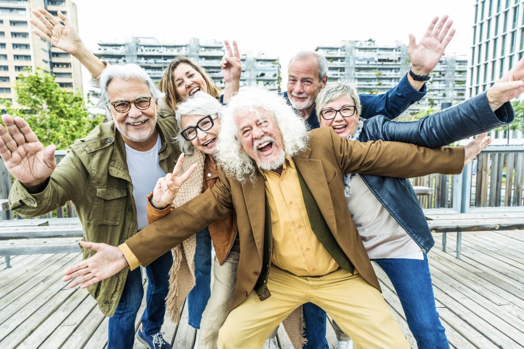 Seis personas adultas saludando y posando para la cámara. Todos ellos felices y entremezclados en una terraza con un fondo de edificios altos.Visten con ropa muy moderna.
