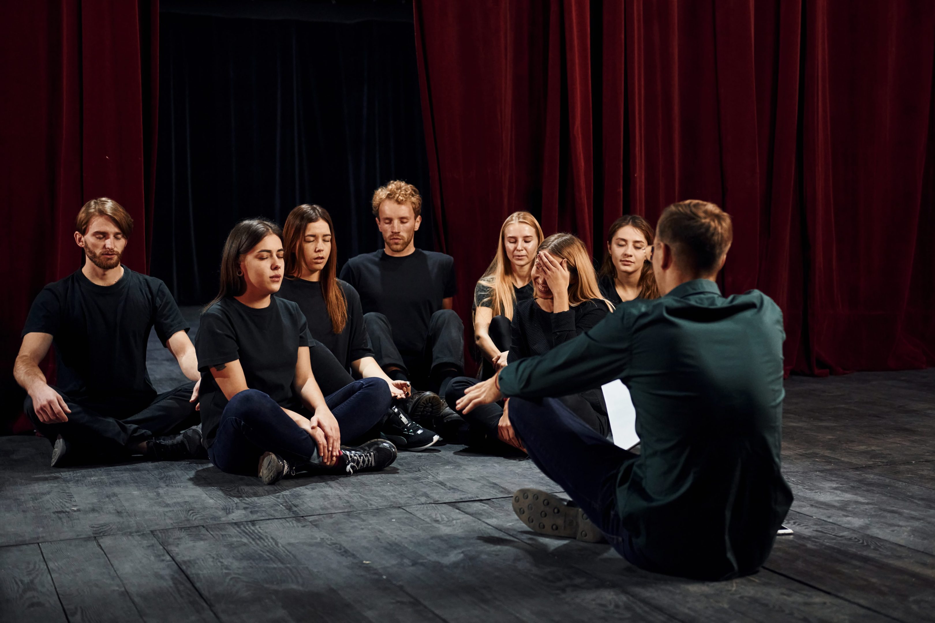 Actores en el escenario en posición de relajación con los ojos cerrados, escuchando las indicaciones del profesor sentando también en el suelo con una hoja en la mano derecha y todos vestidos de negro sobre el fondo negro y unos telones rojos entreabiertos en la mitad