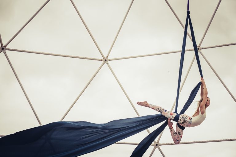 Una chica en el aire con las telas aéreas de color azul y ella con unas mallas de color blanco y cosas estampadas y camiseta blanca de tirantes. Está en un Domo