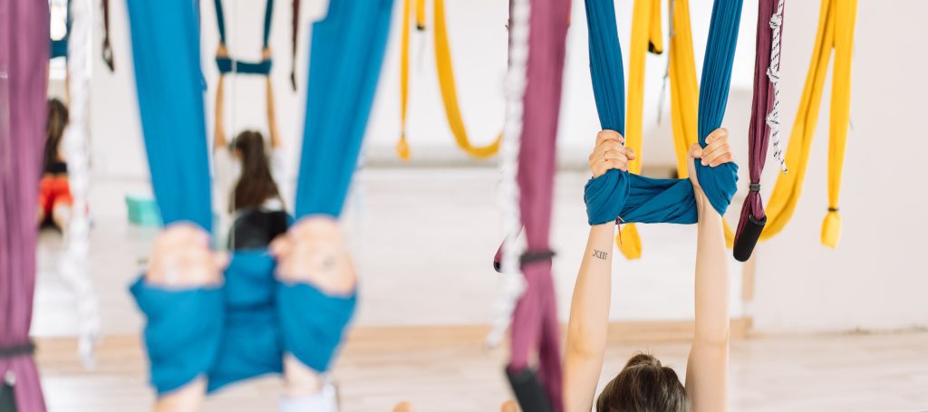 Foto de dos brazos y dos manos cogiendo las telas de aéreo yoga. Las telas que cogen son de color azul, pero se ven telas de color amarillo y morado colgadas en la sala. El fondo refleja las telas por un espejo que hay en la sala.