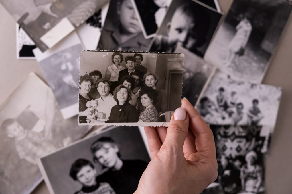 Imagen de una mano sosteniendo fotografía antiguas en blanco y negro con los marcos rebordeados. Muchas más amontonadas y desenfocadas por debajo de la foto que sostiene