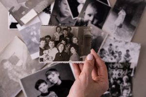 Imagen de una mano sosteniendo fotografía antiguas en blanco y negro con los marcos rebordeados. Muchas más amontonadas y desenfocadas por debajo de la foto que sostiene