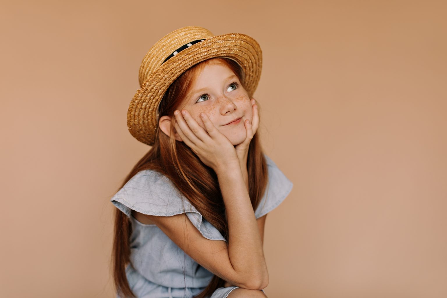 Niña pelirroja posando con vestido azul y sombrero de paja en actitud soñadora