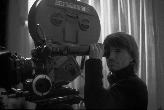 Achero Mañas director de cine mirando a través del visor de una cámara de cine profesional, con una suerter negro de cuello alto y unas cortinas de fondo blanco