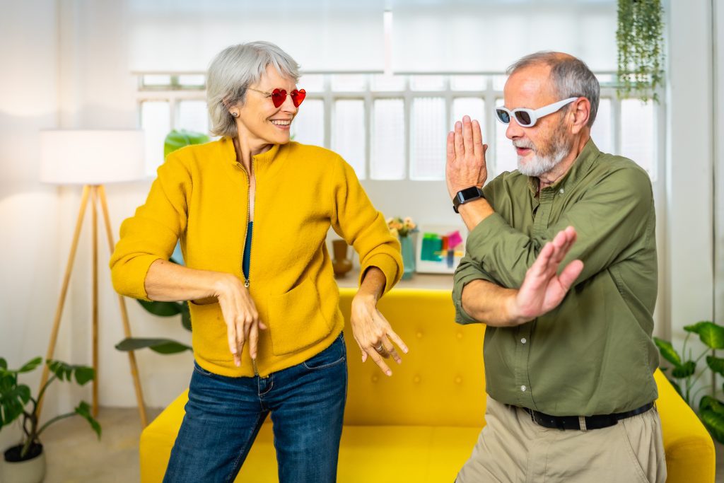Mujer y hombre de la tercera edad, vestidos muy sport y juveniles. Ella viste con un vaquero azul y chaqueta amarilla de cremallera y él con una camisa verde militar arremangada hasta los codos y con un reloj. Están moviendo los brazos como si bailaran y con gafas de sol ambos. Ella lleva gafas en forma de corazón y el blancas y cuadraditas. El fondo un sofá amarillo, unas plantas, una lampara de pie. Parece un set como si fuera un salón