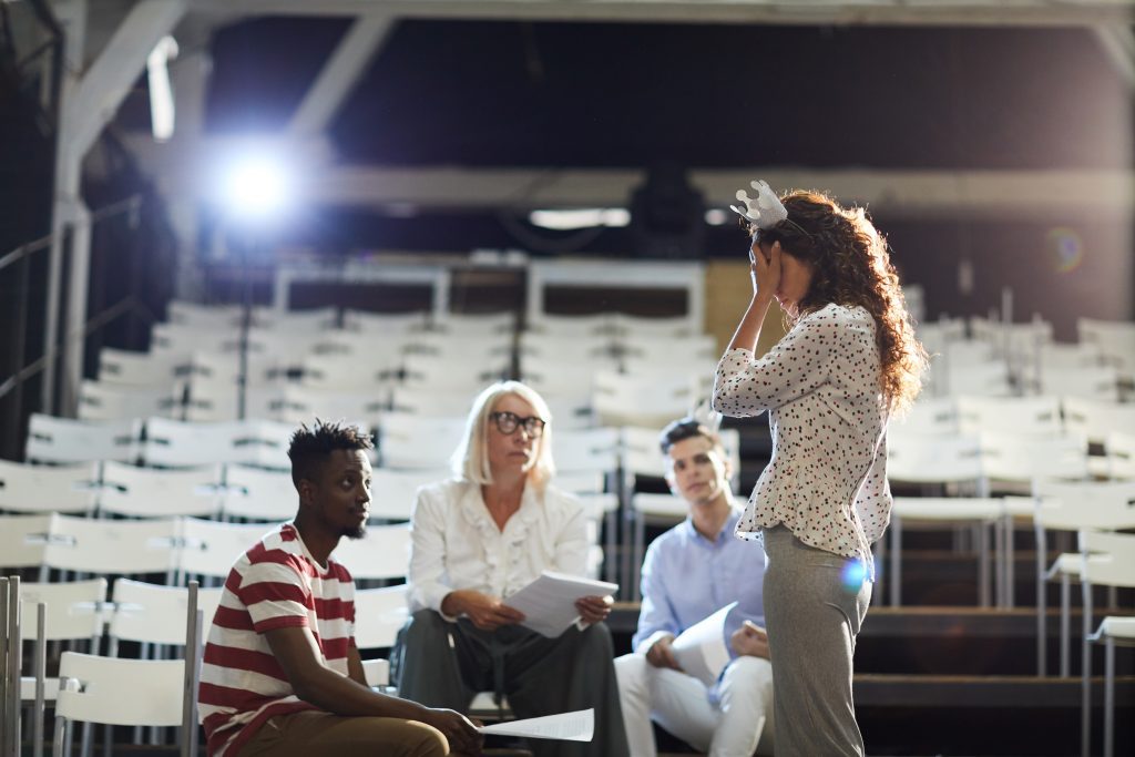 Cuatro actores en unas gragas de teatro con sillas blancas y una luz de fondo blanca que les ilumina. Una de ellas está de pie con una coronita de papel en la cabeza. Los otros tres sentados con los textos en la mano mirando como interpreta con una de las mano llevándosela a la cabeza.