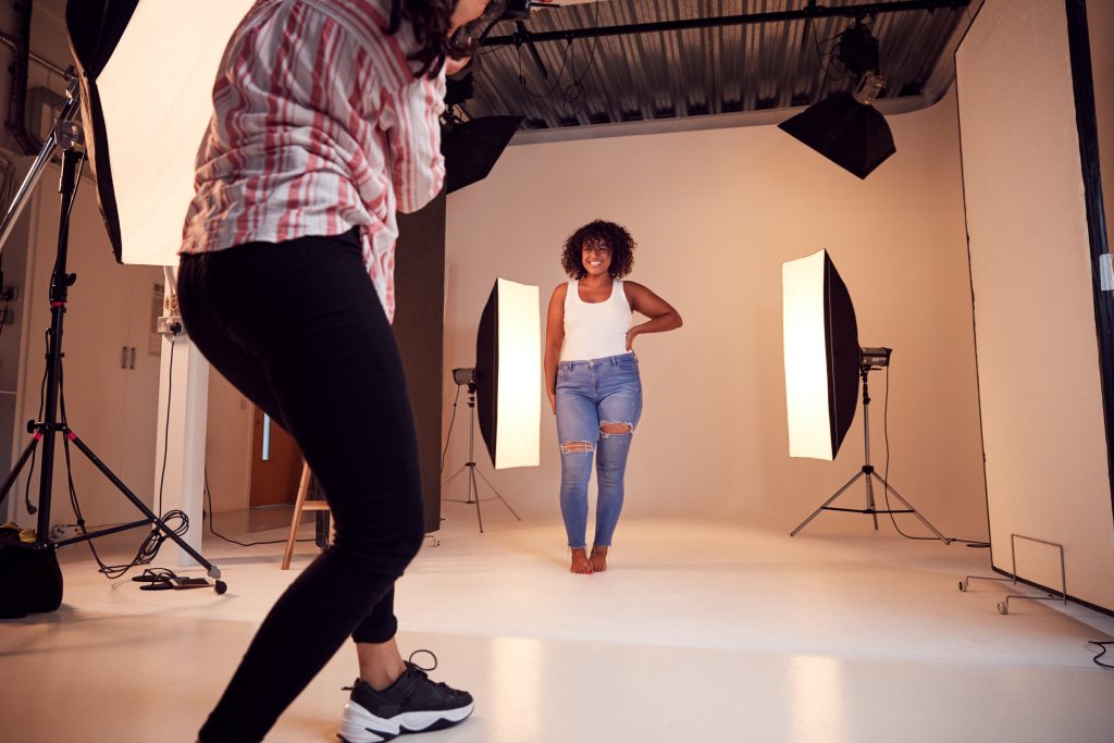 Fotógrafa y modelo en un estudio. Fondo blanco, tejas de color negro. Dos luces colgadas en las varas y dos más junto a sus trípodes en los laterales de la modelo y otra luz frontal al lado de la fotógrafa. La modelo lleva una camiseta de tirantes blancos y la fotógrafa lleva una camisa a rayas rosas y un pantalón negro. La modelo va descalza y la fotógrafa va con zapatillas de deporte negras.