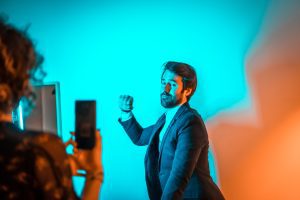 Un chico con traje de chaqueta posando con el brazo cerrando el puño como señal de lucha obrera, y una chica haciendo sosteniendo un móvil con ambas manos. Fondo azul claro.