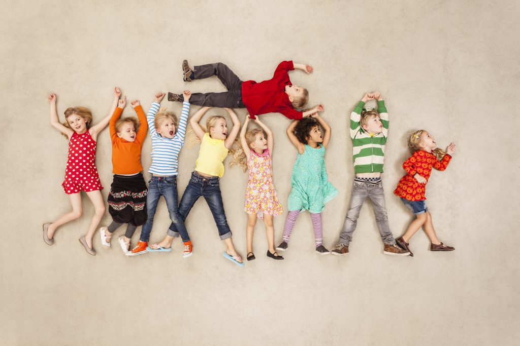 Grupo de 9 niños tirados en el suelo de la sala juntos con las manos hacia arriba. Uno de ellos está encima de las manos de 4 de ellos. Están como volviendo de la relajación