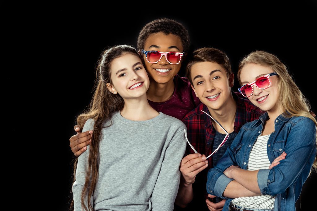 Cuatro chicos sonriendo a cámara. Tres de ellos llevan gafas de color rosa y uno de ellos la sostiene en sus manos. Una de las chicas no lleva ni coge las gafas. Están posando para la cámara, el fondo es negro.