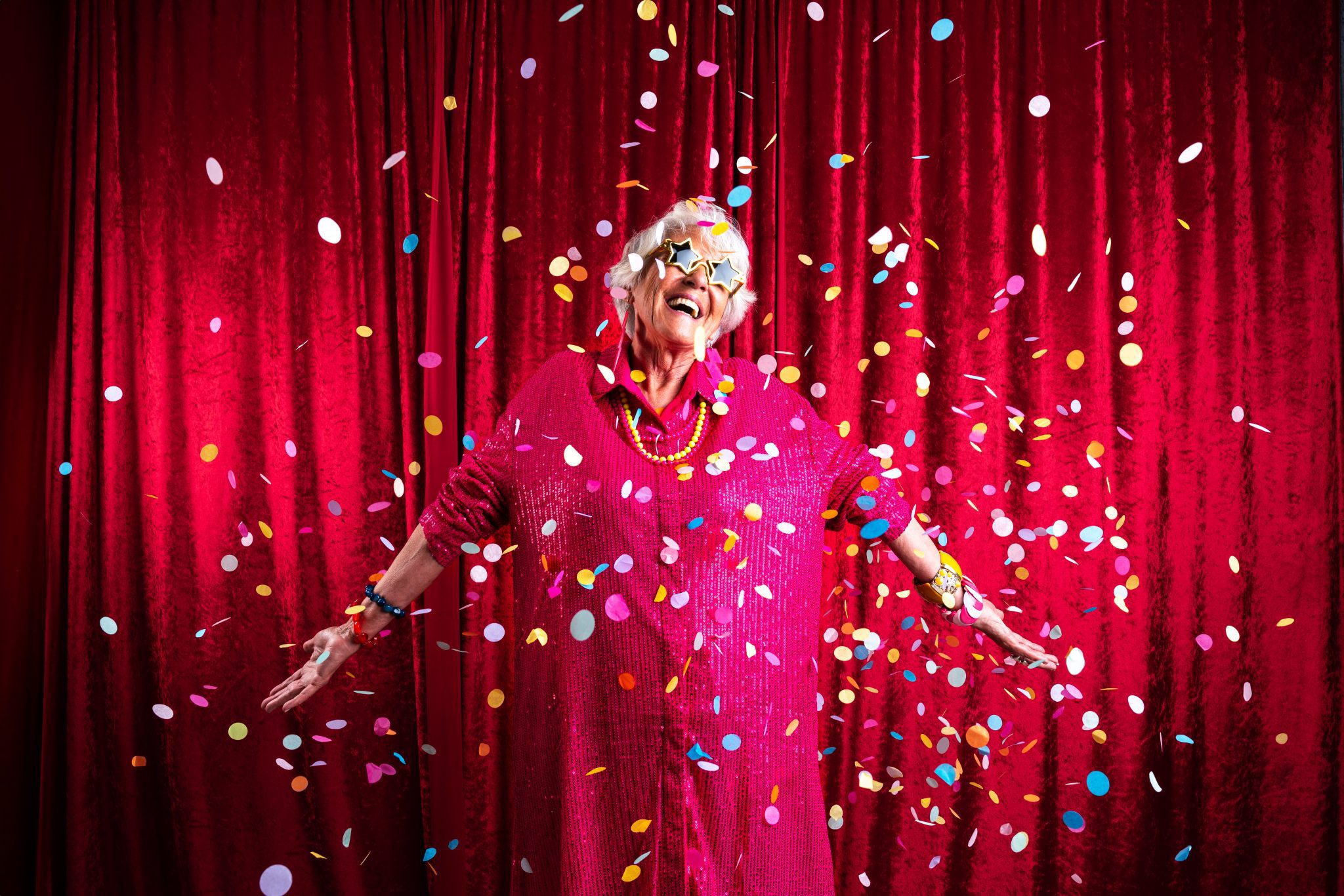 Mujer con vestido color morado y sobre fondo rojo, con gafas en forma de estríela y con pulseras de diferentes colores en las muñecas y un colgante amarillo. Están cayendo del cielo Confeti de diferentes colores y los recibe con los brazos abiertos y con signos de alegría y felicidad.